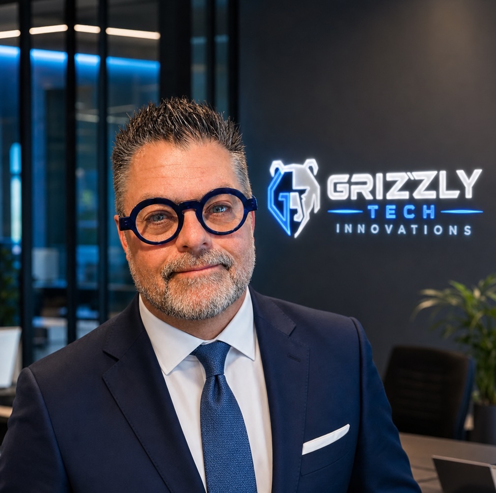 Portrait of a smiling man in a navy suit and blue tie, wearing round blue glasses, in an office with a Grizzly Tech Innovations sign in the background.